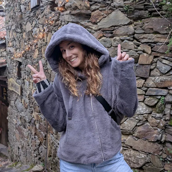Smiling in a grey hooded fleece and making peace signs in front of an old stone wall.