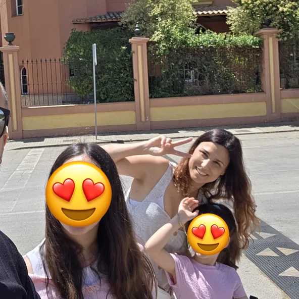 A sunny family moment on a street: smiling and making a peace sign, with two children whose faces are covered by heart-eyes emoji.